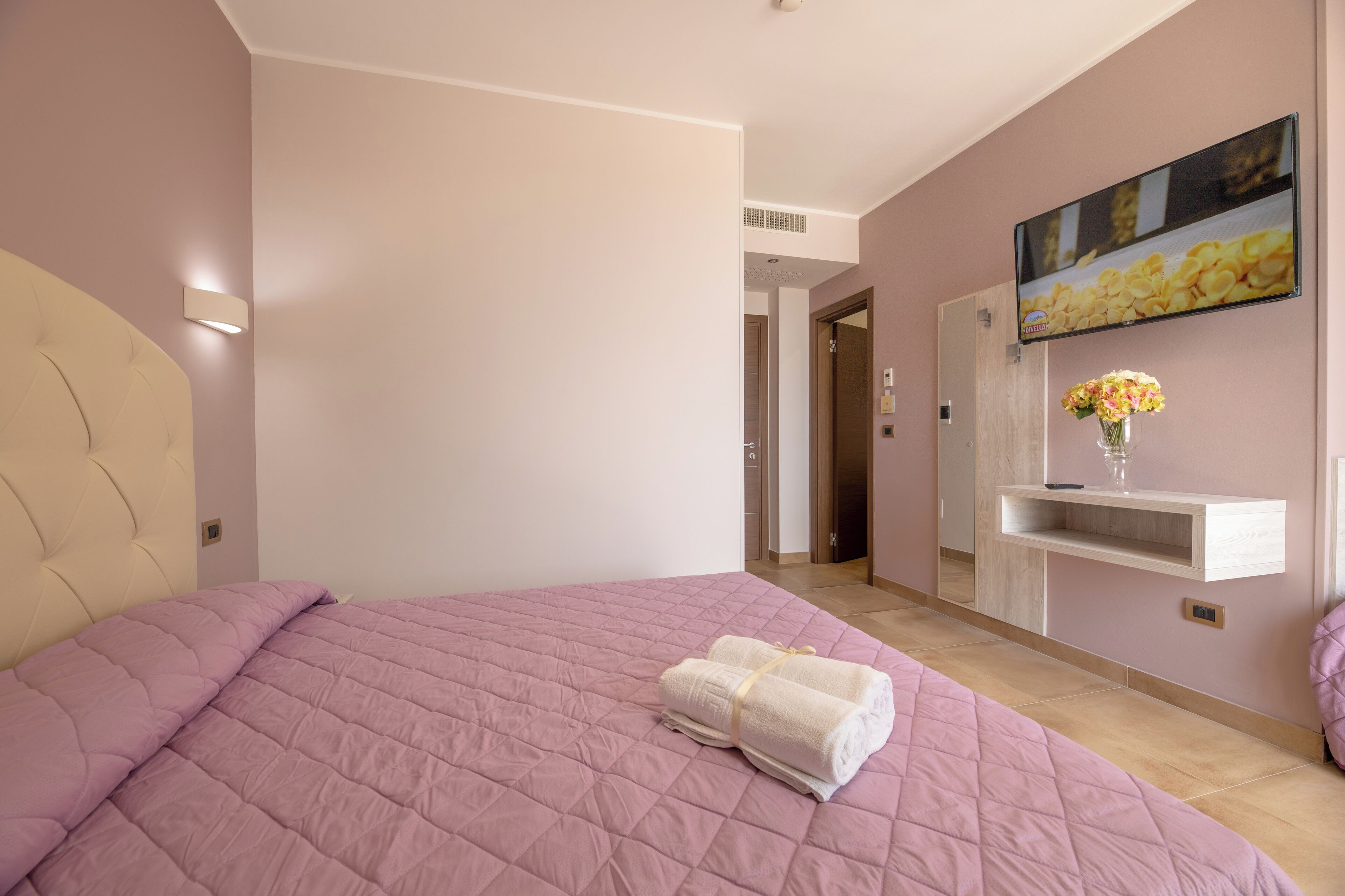 A bedroom with a pink quilted bedspread, a neatly made white towel, a wall-mounted TV, and a small vase with flowers on a shelf.