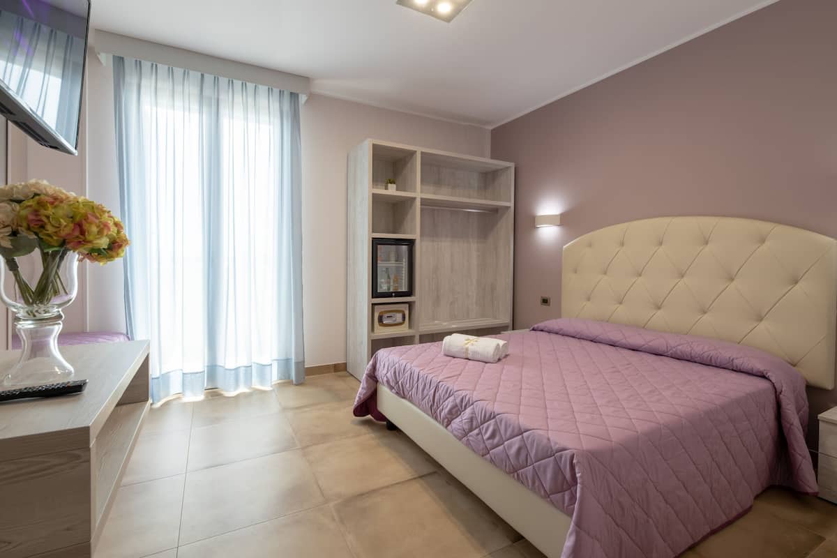 Un dormitorio con cama, tocador con espejo, televisor, armario y ventana con cortinas.