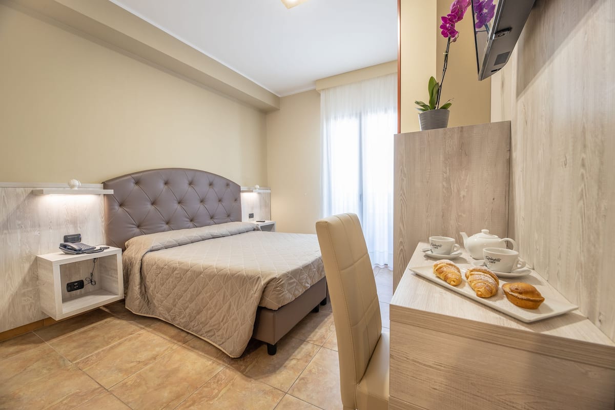 Una habitación de hotel con cama, mesitas de noche, un escritorio y una bandeja con pasteles, y un televisor en la pared.