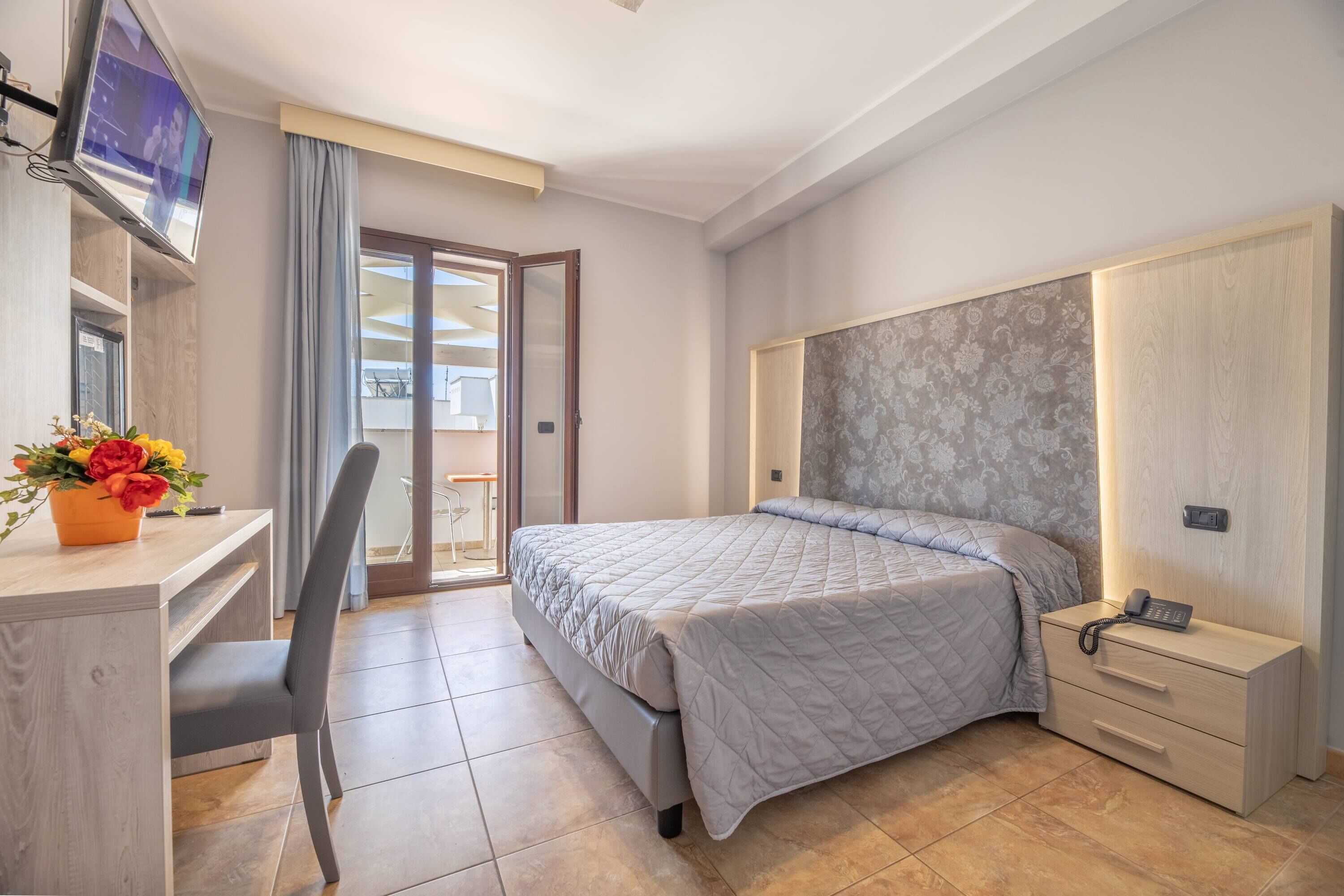 Een moderne hotelkamer met een bed, een bureau met stoel, een televisie en een kleintje tafel met een bloevas.
