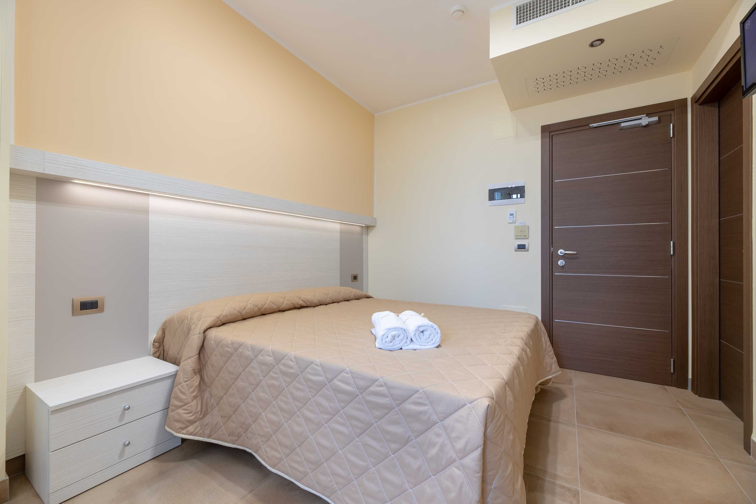 Una cama bien hecha con cubrecama acolchado y toallas enrolladas, una mesita de noche blanca con cajones, un armario de madera y una lámpara en el techo.