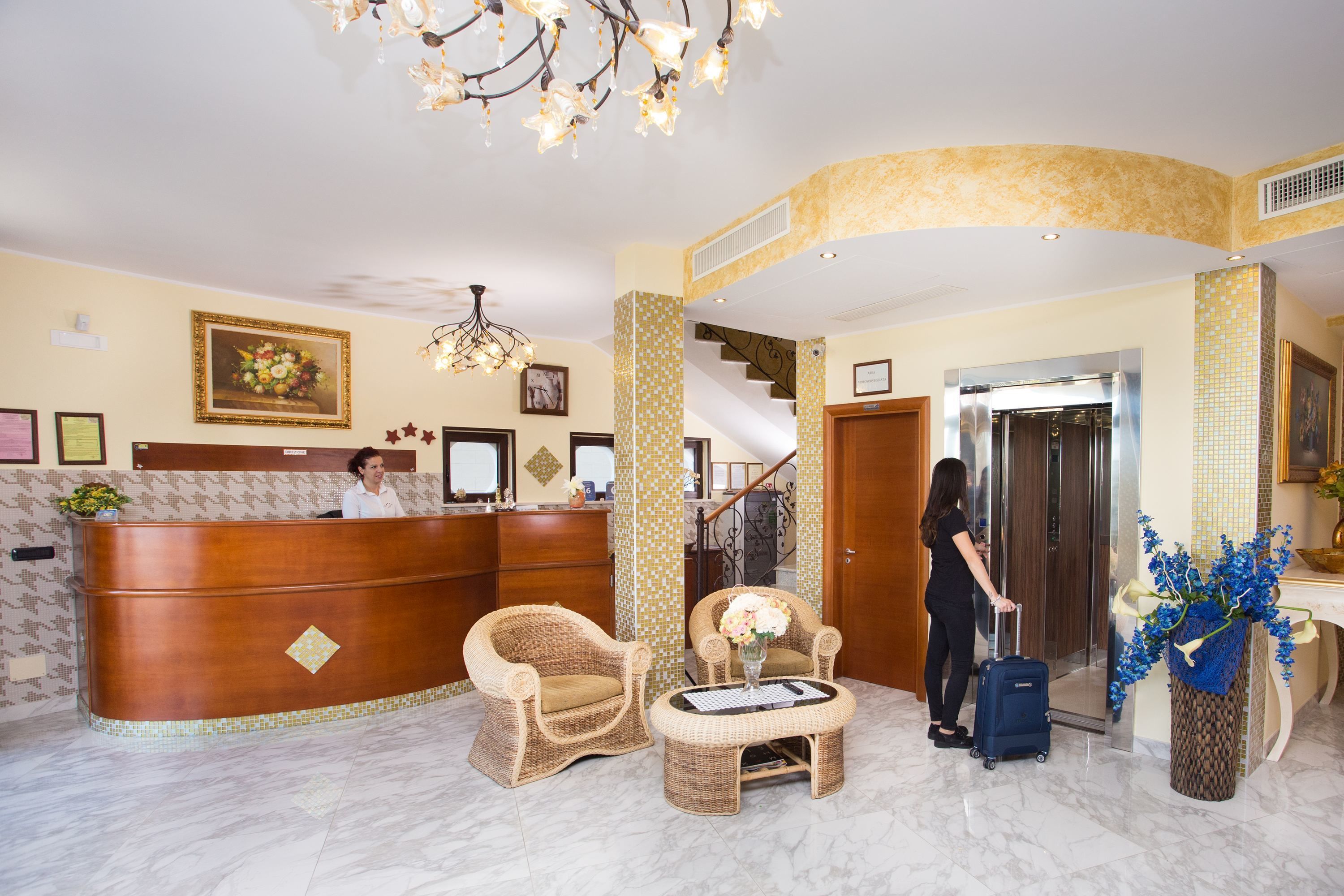 A reception area with a wooden reception desk, wicker chairs, a small table with a vase, and a blue suitcase.