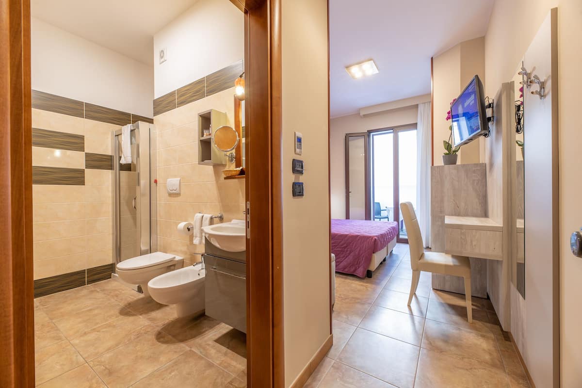 Un baño moderno con ducha, inodoro y lavamanos. Hay un espejo sobre el lavamanos y un televisor montado en la pared.