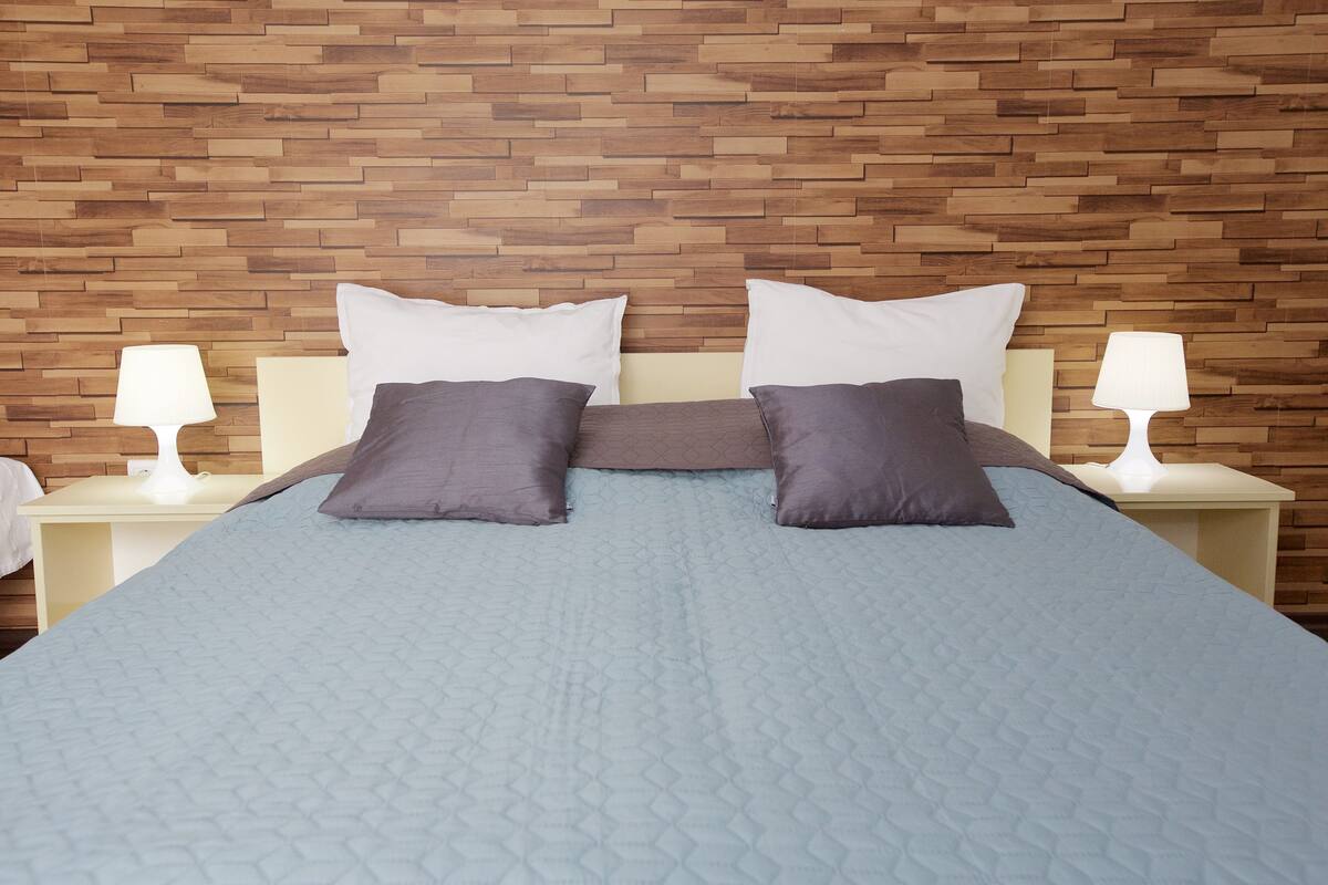 Una cama bien hecha con un edredón azul, dos almohadas blancas y dos almohadas gris oscuro. Dos lámparas de noche sobre mesitas blancas. La pared detrás de la cama está hecha de tablones de madera.