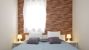A neatly made bed with a quilted blue bedspread, two pillows, and a headboard with a wooden pattern. There are two bedside tables with lamps on either side of the bed. - Sunset Apartments (Hajduszoboszlo)