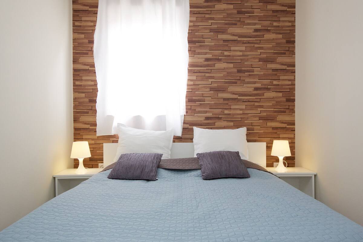 Una cama bien hecha con una colcha azul acolchada, dos almohadas y un cabecero con diseño de madera. Hay dos mesitas de noche con lámparas a cada lado de la cama.