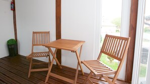 A wooden table and two chairs on a wooden deck with a white wall and a window. - Sunset Apartments (Hajduszoboszlo)