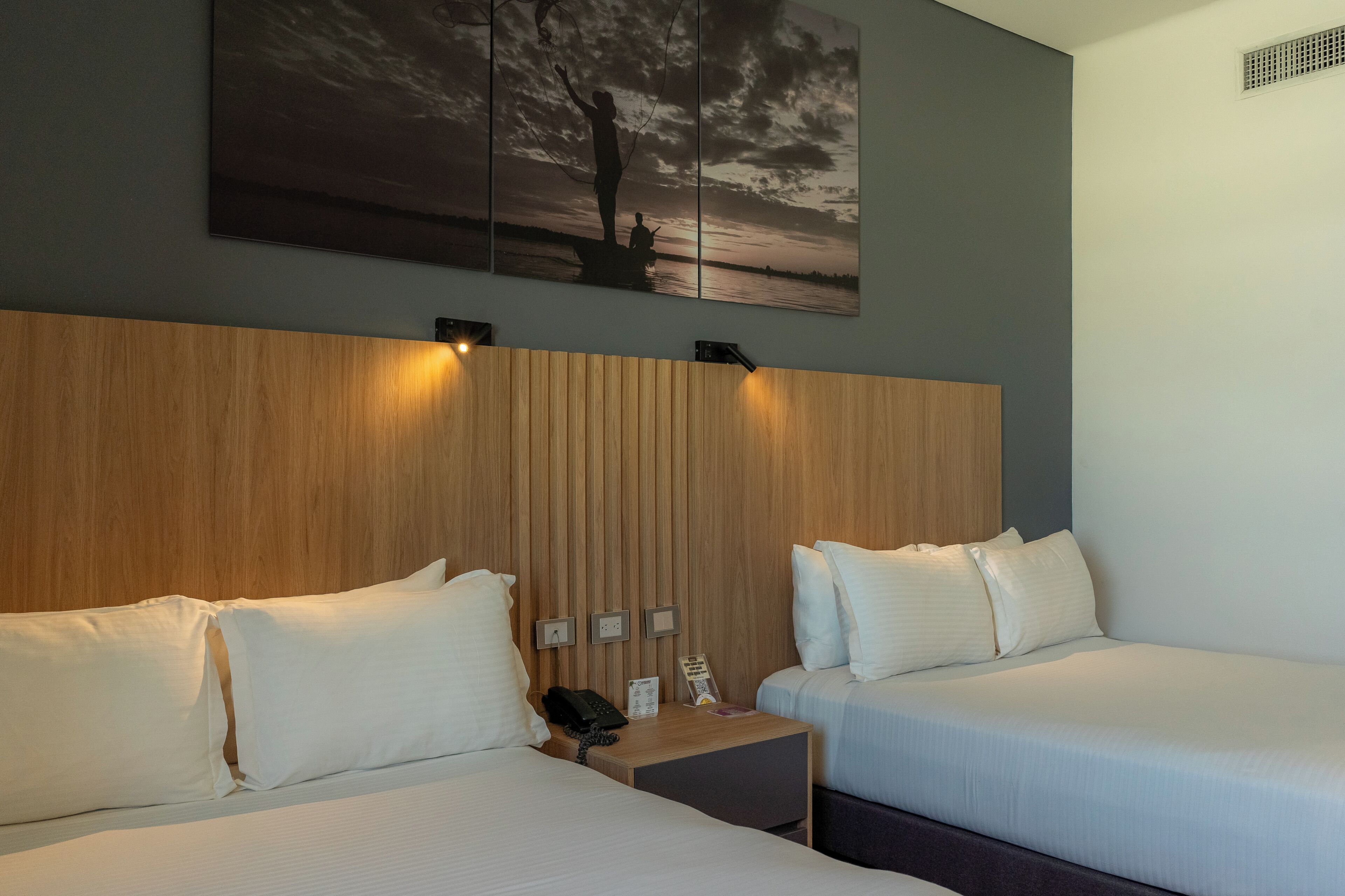 Habitación de hotel con dos camas, cabecera de madera, mesita de noche con teléfono y un cuadro de un atardecer.