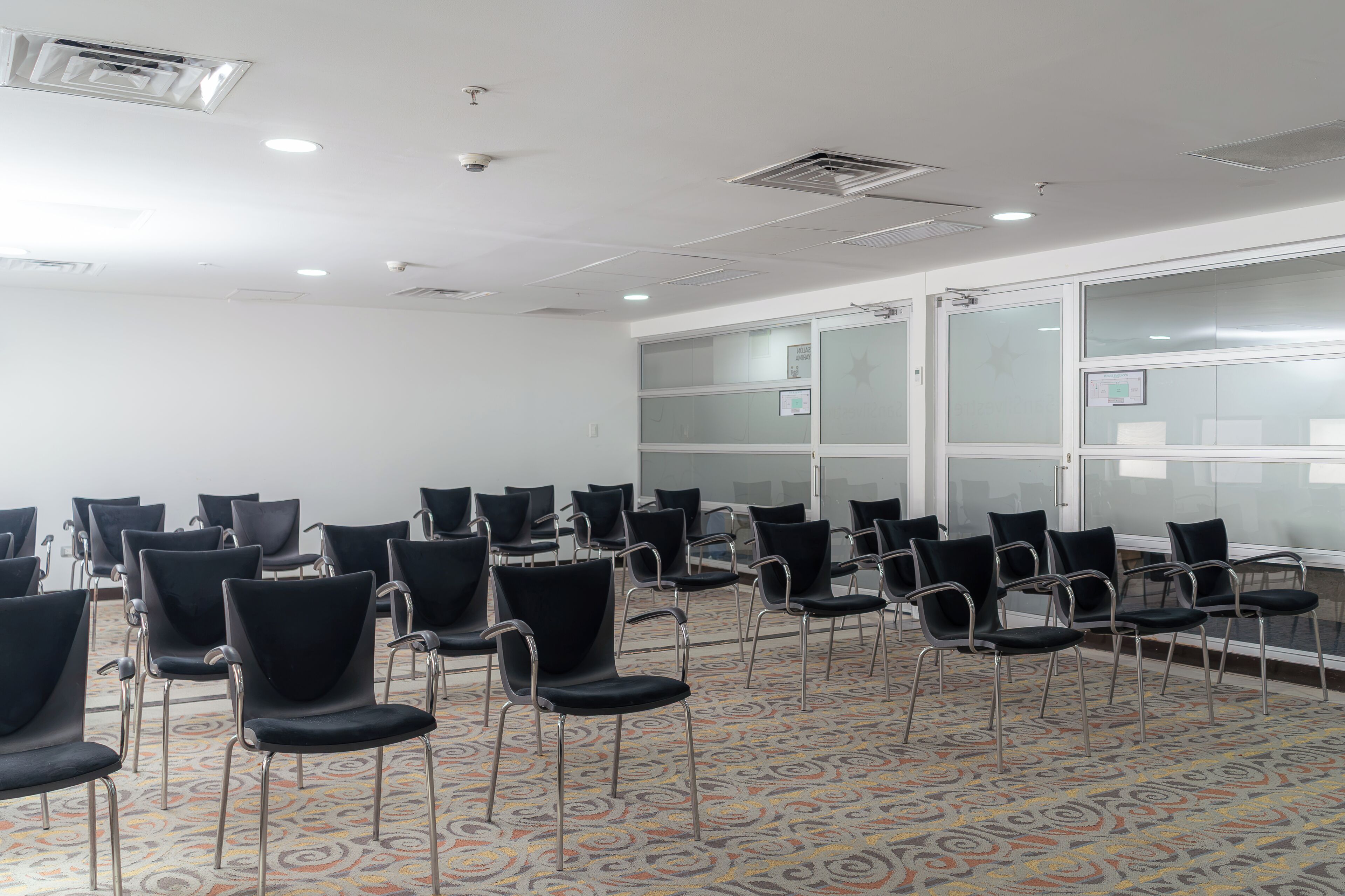 A conference room with rows of chairs, a patterned carpet, and a white ceiling with recessed lighting.