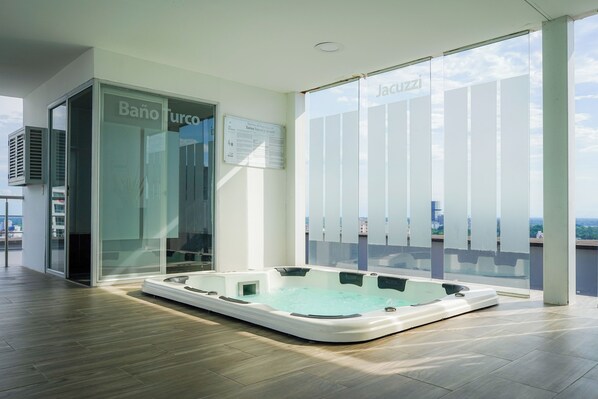 A modern hotel room with a glass door entrance, a large window with frosted glass panels, and a hot tub in the center.