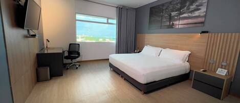 A modern hotel room with a large bed, a desk, a chair, a TV, and a window with a view.