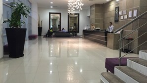 A modern hotel lobby with a reception desk, a large potted plant, a staircase, and a chandelier.