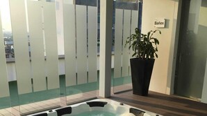 A hot tub on a wooden deck with a glass partition and a potted plant nearby.
