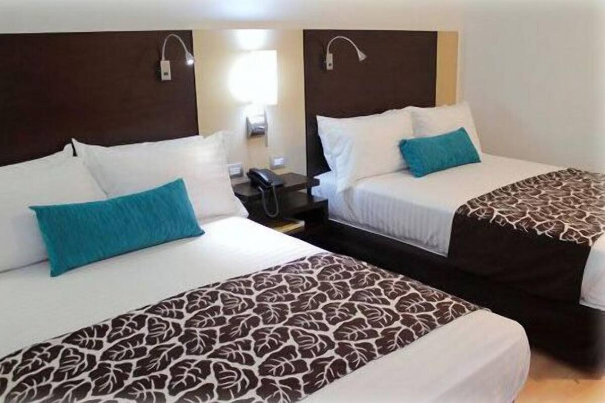 Habitación de hotel con dos camas individuales, cada una con una colcha estampada y un cojín verde azulado. Entre las camas, un mesita de noche con un teléfono y una lámpara.