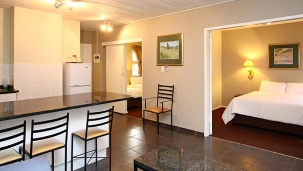 A compact hotel room with a kitchenette, a dining area with chairs, a bed, and a nightstand with a lamp. - Vetho 2 Apartments OR Tambo Airport (Kempton Park)