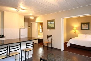 A compact hotel room with a kitchenette, a dining area with chairs, a bed, and a nightstand with a lamp. - Vetho 2 Apartments OR Tambo Airport (Kempton Park)