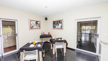 A dining area with three tables, each with a black tablecloth and white chairs. There are framed pictures on the walls, and a small table with a fruit bowl and drinks.