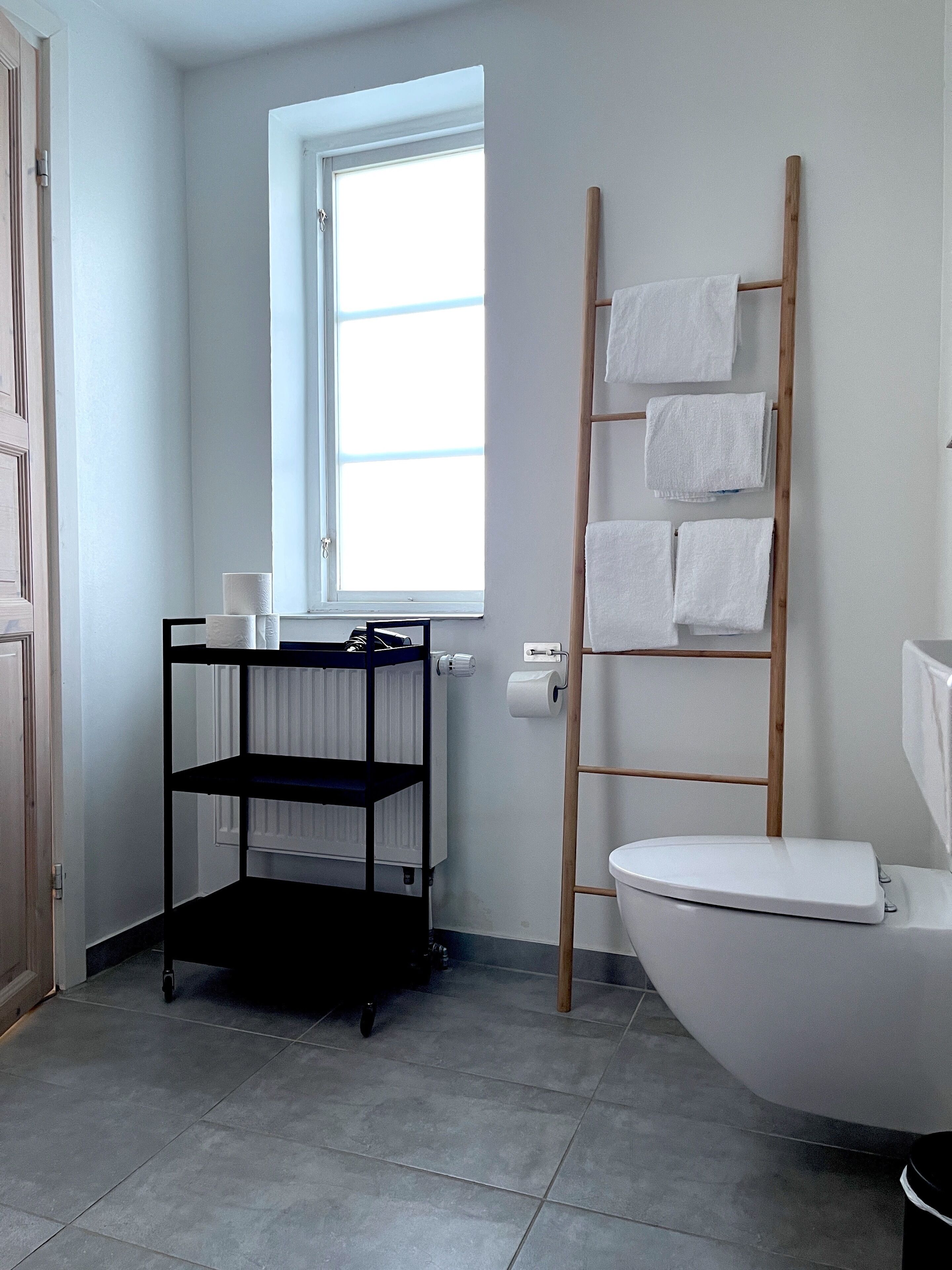 Un baño con un inodoro, una escalera de madera con toallas, un estante negro y una ventana.