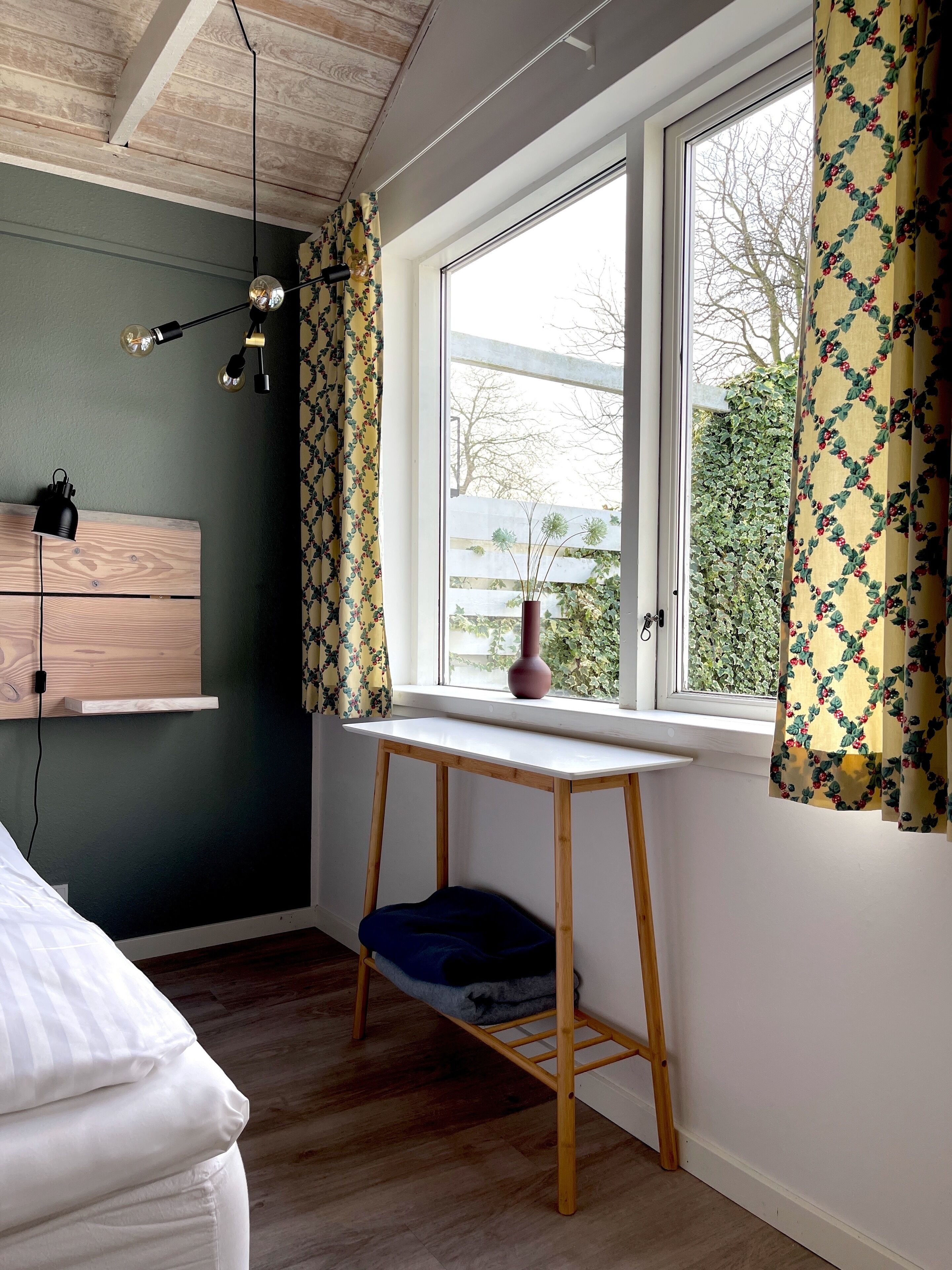 Una habitación con una cama, una mesita con un florero, una ventana con cortinas y un techo de madera.