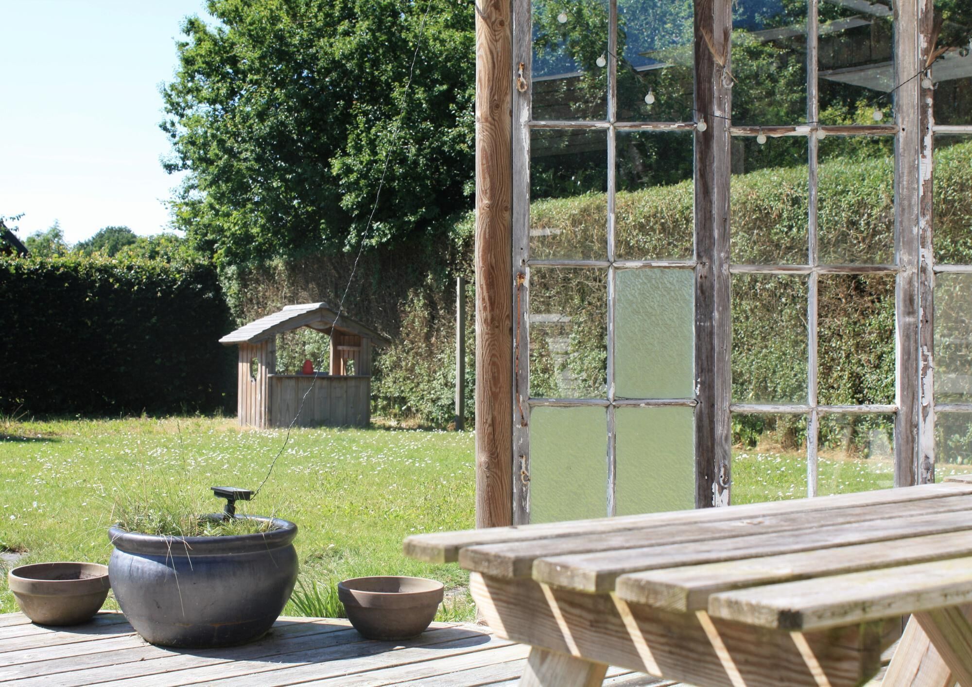 A wooden deck with a rustic table and two bowls, overlooking a grassy area with a small wooden structure and a pond.