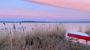 Escena de playa con pastos altos, un paraguas rojo y blanco, y una masa de agua tranquila bajo un cielo rosa y azul.
