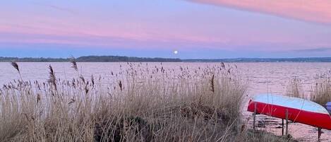 Escena de playa con pastos altos, un paraguas rojo y blanco, y una masa de agua tranquila bajo un cielo rosa y azul.