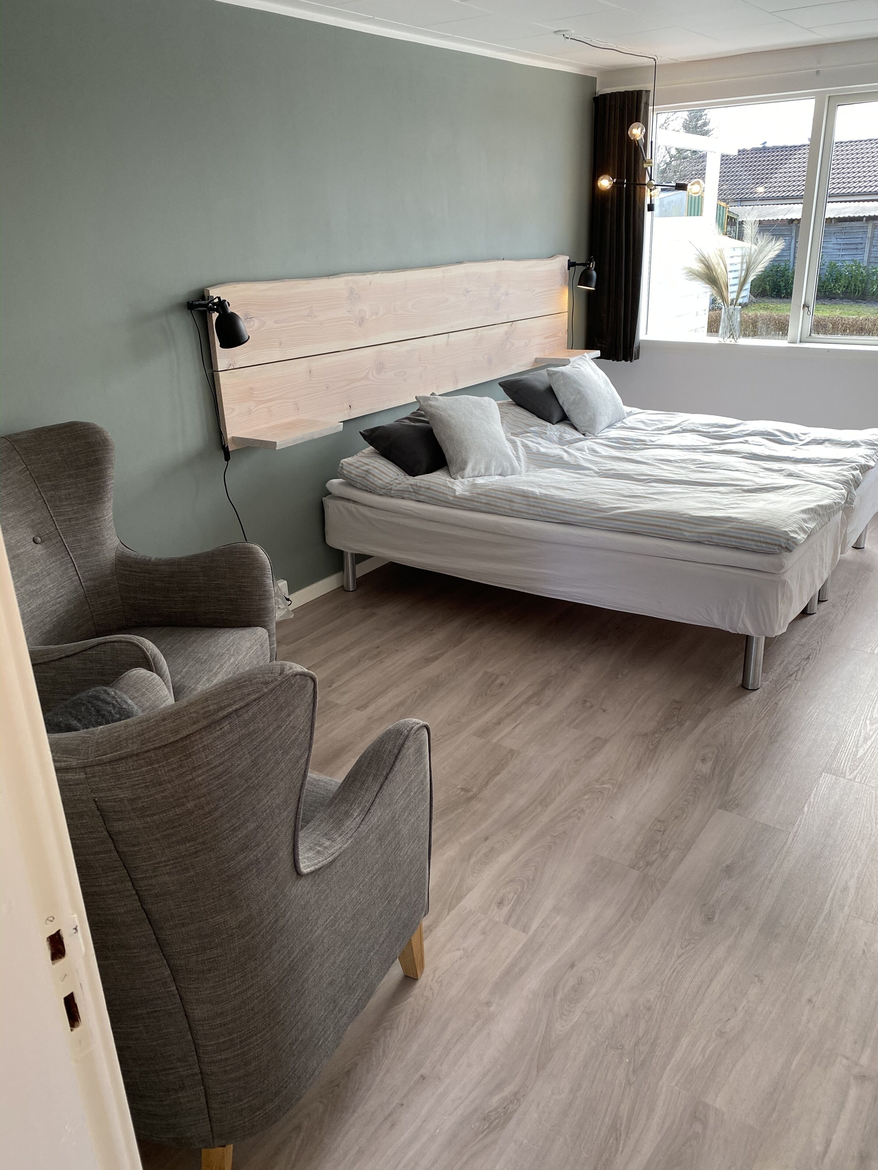 A bedroom with a bed, two chairs, a wooden headboard, and a window with curtains.