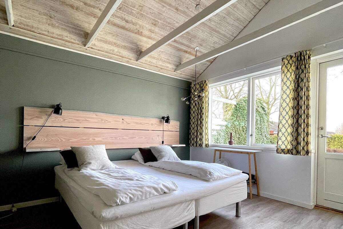 Une chambre avec un plafond en bois, un lit avec une literie blanche, une tête de lit et une fenêtre avec des rideaux.