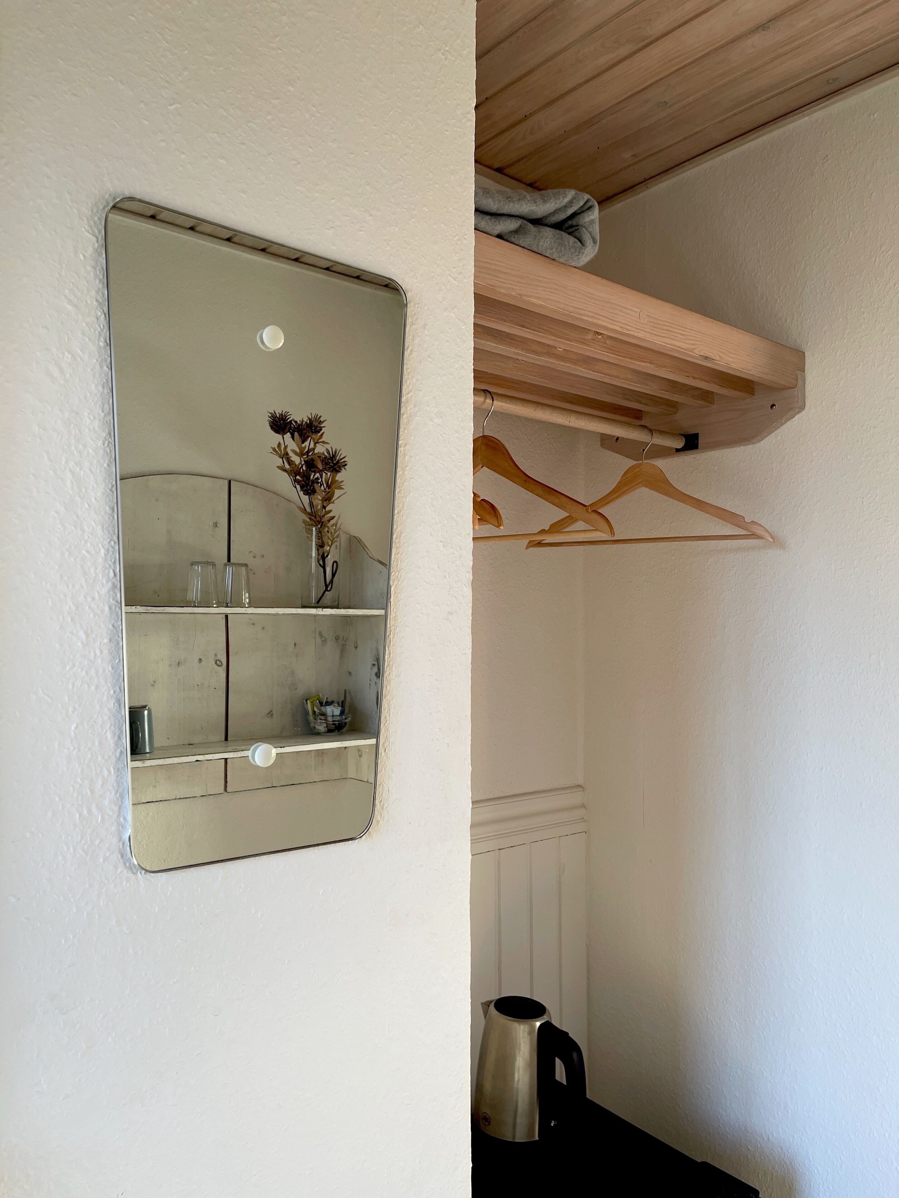 A mirror reflecting a bathroom vanity with a vase of dried flowers, a kettle, and a shelf with various items.