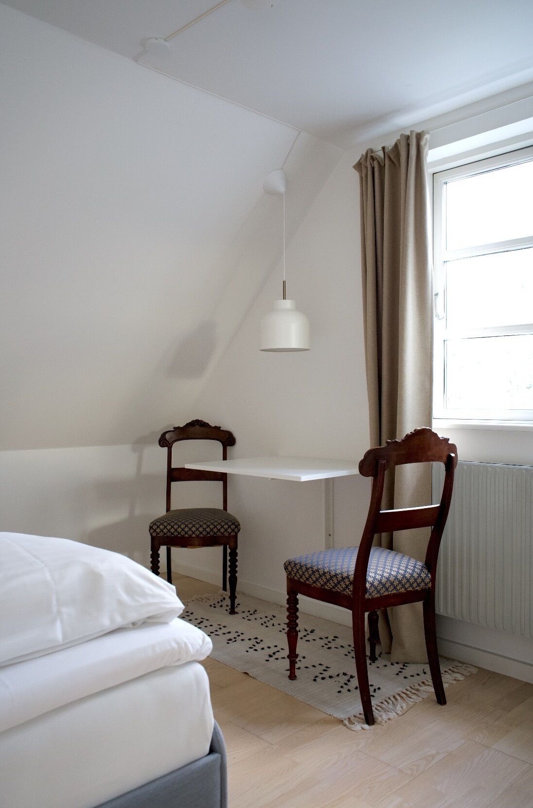 Una habitación pequeña y bien iluminada con una cama, dos sillas de madera, una mesa blanca y un ventanal con cortinas.