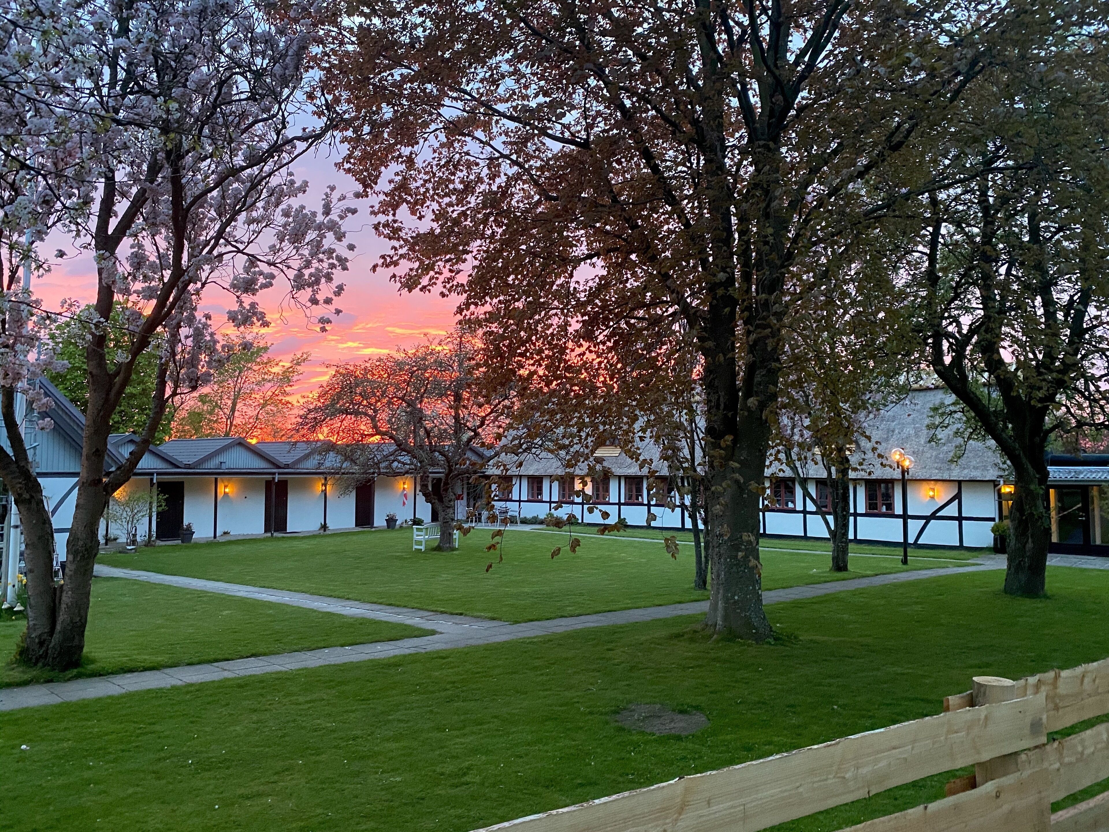 A courtyard with a pathway leading to a row of traditional-style buildings, surrounded by trees and a sunset sky.