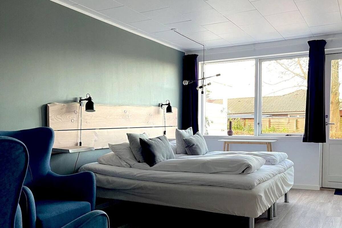 Une chambre d’hôtel moderne avec un grand lit, un fauteuil bleu et une vue sur l’extérieur grâce à une grande fenêtre.