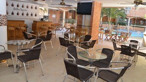 Un espace café en plein air avec des chaises en osier et des tables en verre, un téléviseur fixé au mur, et une piscine visible au fond.