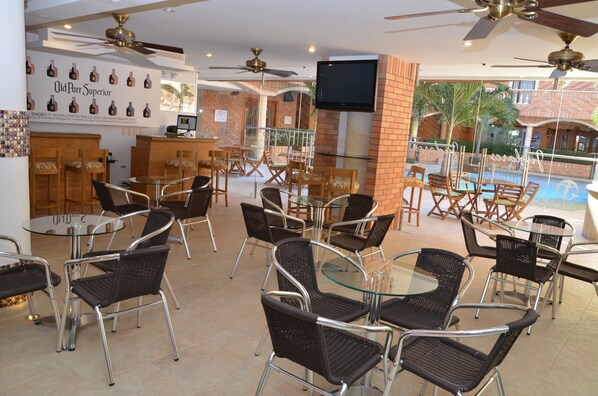 An outdoor cafe area with wicker chairs and glass tables, a TV mounted on the wall, and a pool visible in the background. - Tativan Hotel (Valledupar)