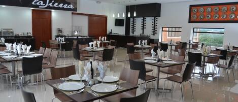 Restaurante com várias mesas preparadas para refeições, área de cozinha e um grande painel fixado na parede.