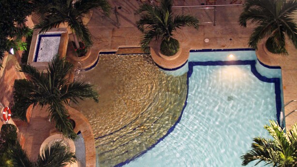 Vue aérienne d’une piscine dotée d’un îlot circulaire, entourée de palmiers et de chaises longues.