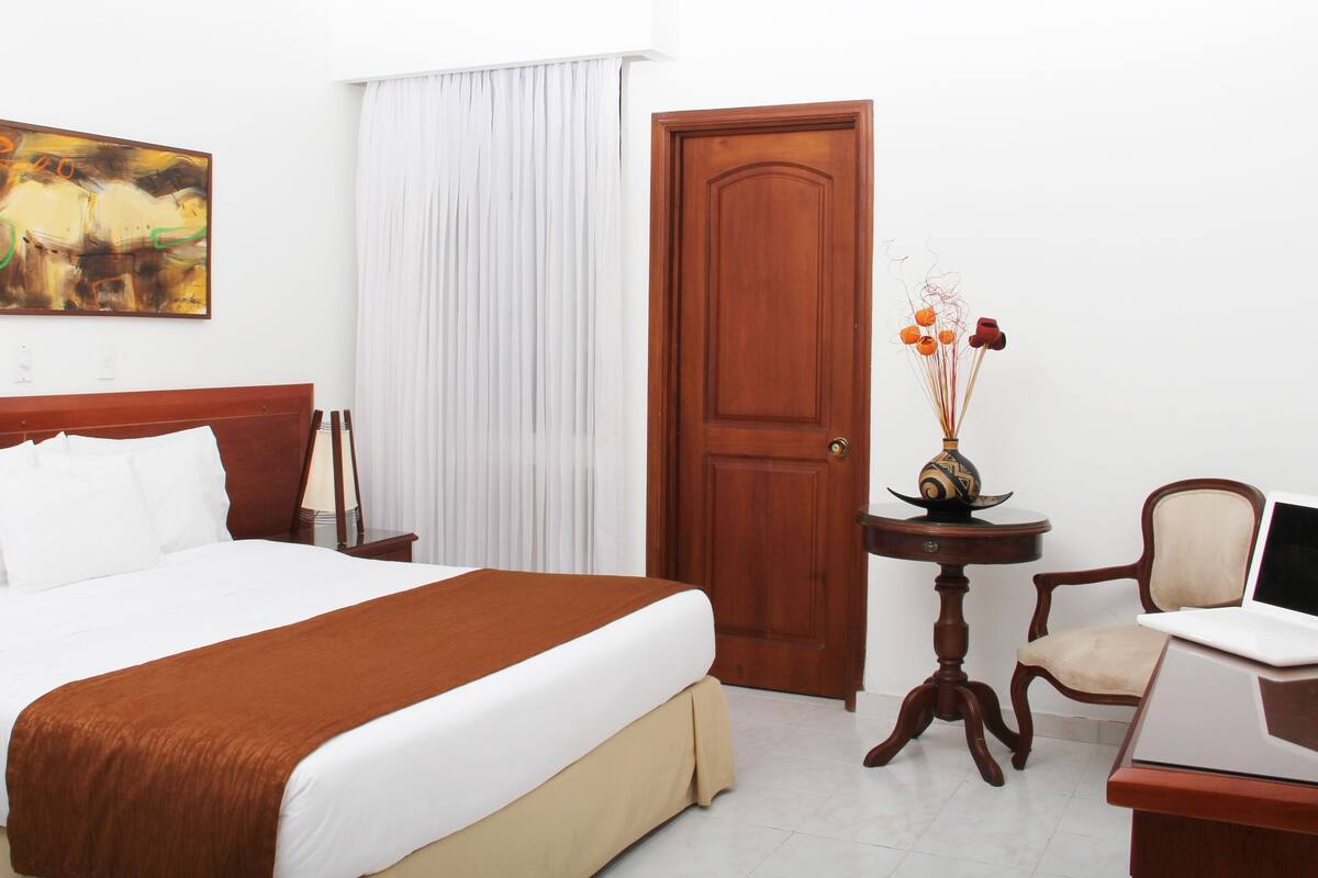 Un cuarto de hotel con una cama grande, cabecera de madera, un escritorio con una silla, una mesita con un florero y un cuadro en la pared.
