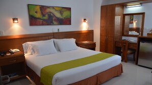Une chambre d’hôtel avec un lit, des tables de chevet, un bureau, une chaise, un miroir et un tableau coloré accroché au mur.