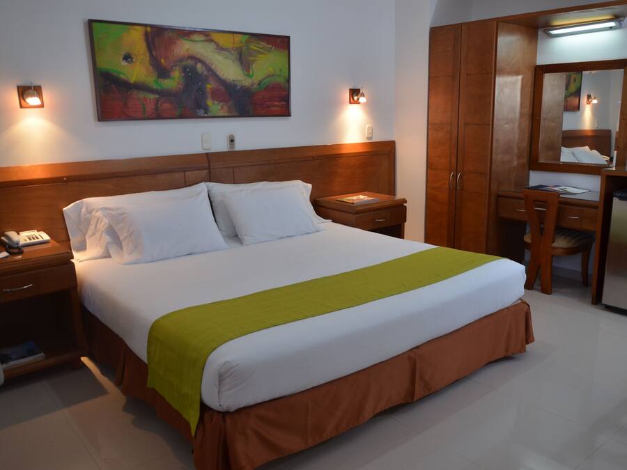 A hotel room with a bed, bedside tables, a desk, a chair, a mirror, and a colorful painting on the wall.