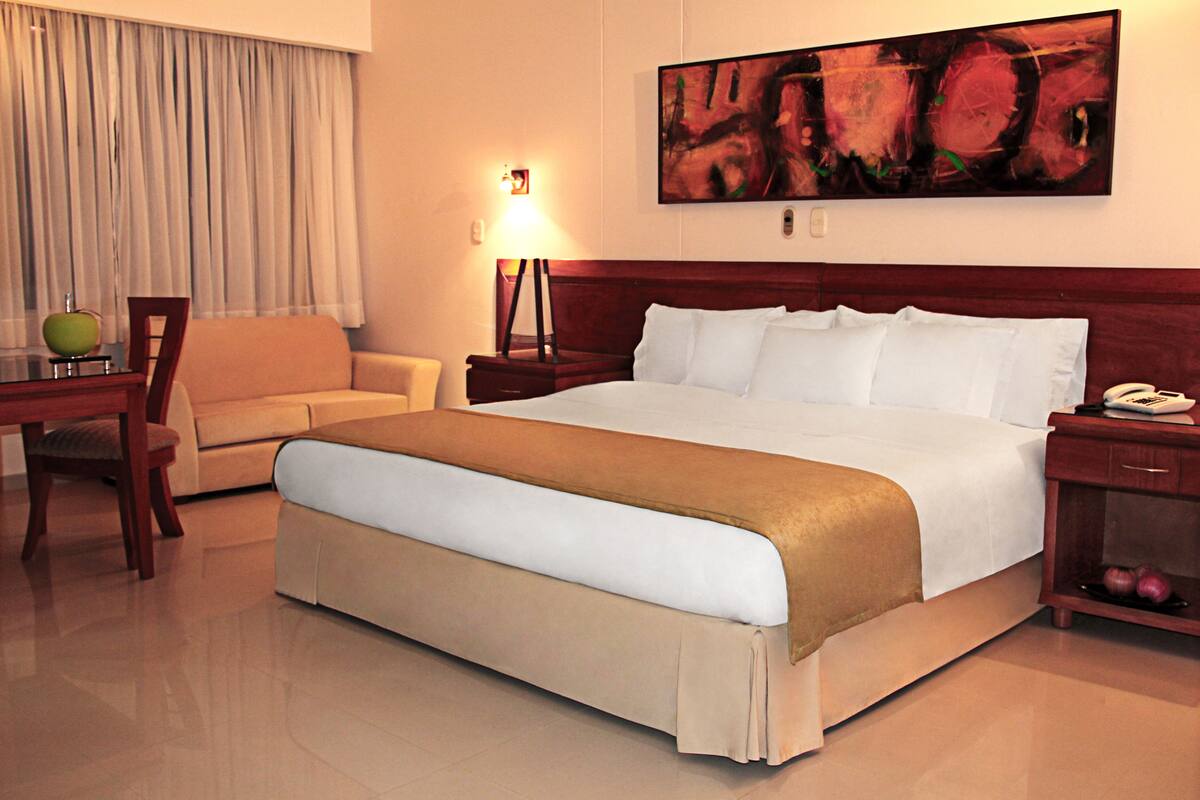 Habitación de hotel con una cama grande, un escritorio con lámpara, una silla y un cuadro en la pared.