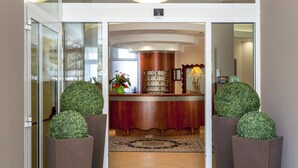 Un espace d’accueil avec un comptoir en bois, un grand miroir et un tapis décoratif. Deux grands plantes en pot sont placées de part et d’autre du comptoir.