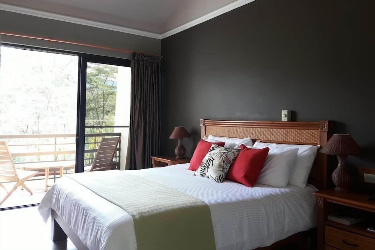 Un dormitorio con cama, mesitas de noche, lámpara, ventilador de techo, ventana con cortinas y balcón con sillas.