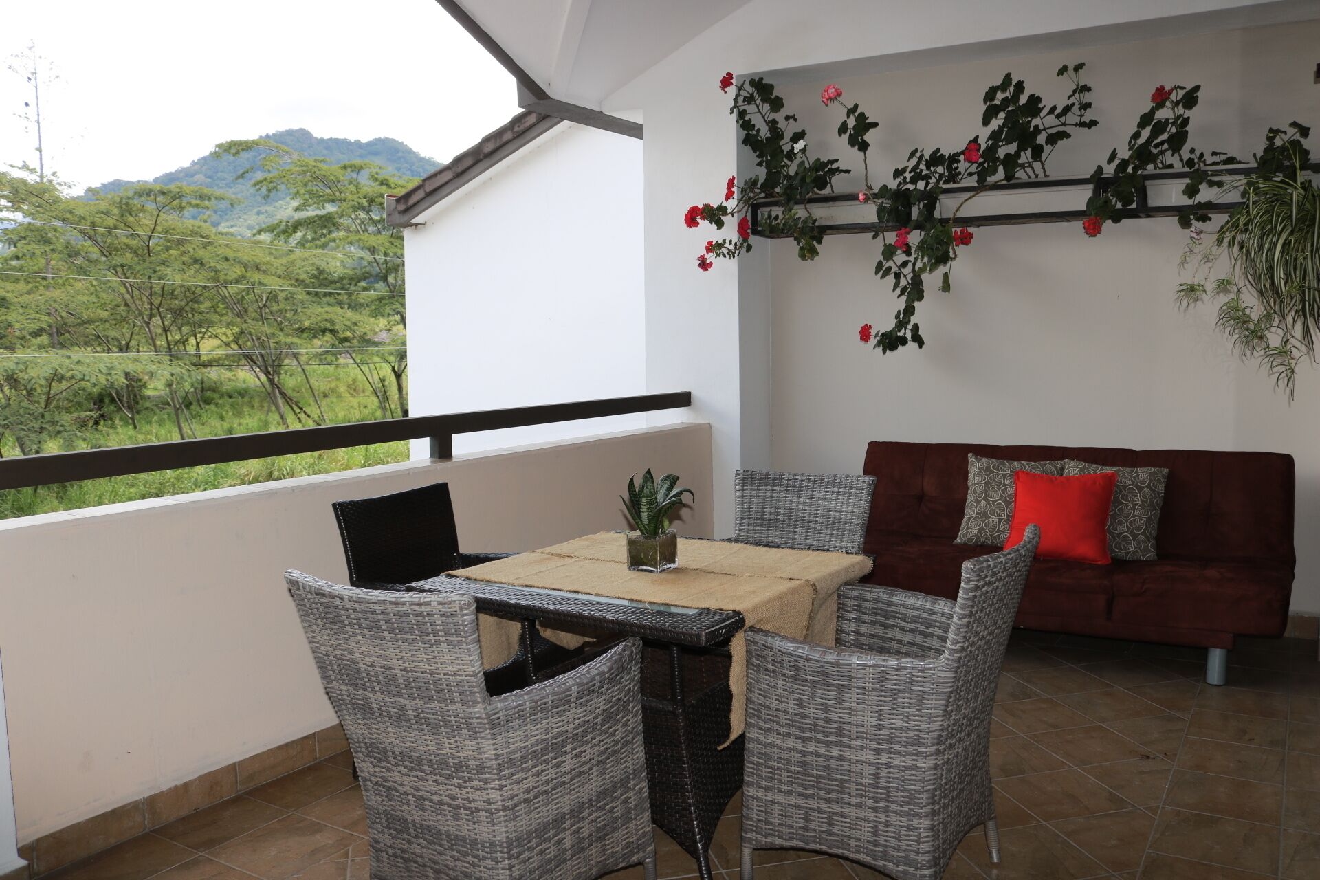 A balcony with wicker furniture, a table, and a sofa with red cushions. There is a potted plant on the table and a wall-mounted plant with red flowers.