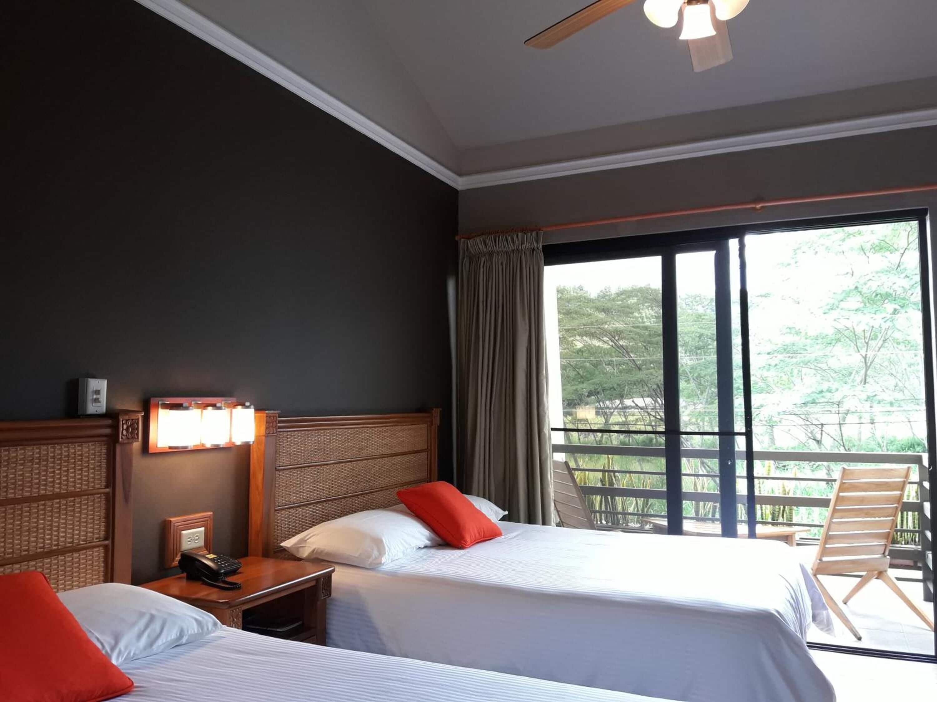 Habitación de hotel con dos camas, una mesita de noche con teléfono, una lámpara de pared y un ventanal con vistas a zonas verdes.
