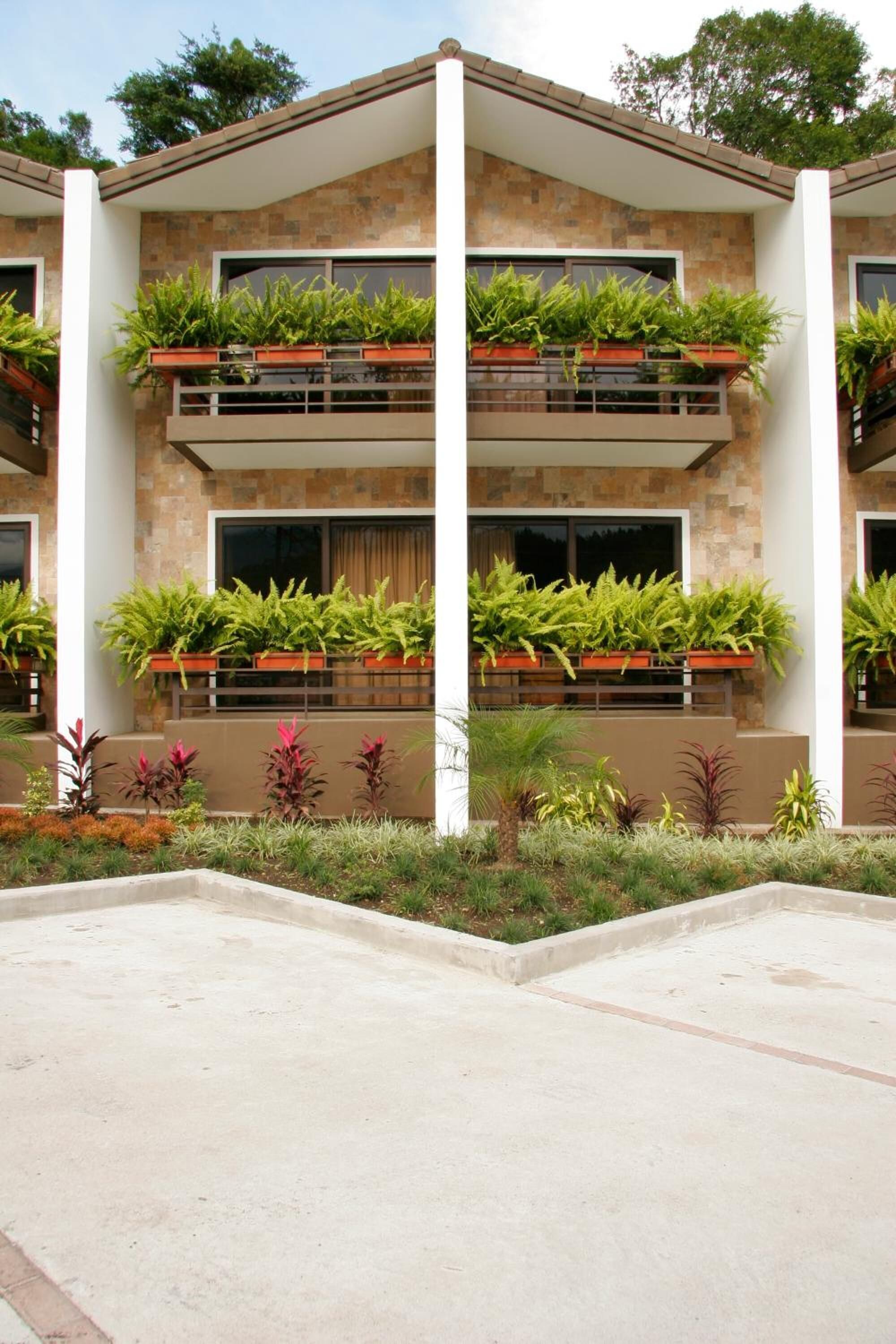 A modern building with a flat roof, two balconies with potted plants, and a concrete ground area.