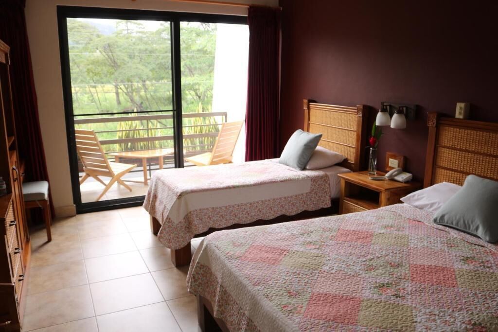 A hotel room with two beds, a balcony with a table and chairs, a wooden nightstand with a lamp, and a view of greenery outside.
