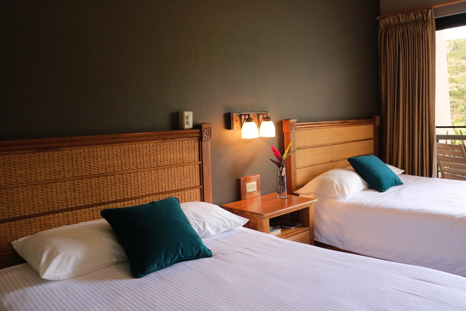 Two beds with headboards, a nightstand with a lamp, and a window with curtains.