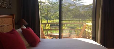 Ein Bett mit roten Kissen, einem hölzernen Kopfteil und Blick auf einen Balkon mit Gartenmöbeln.