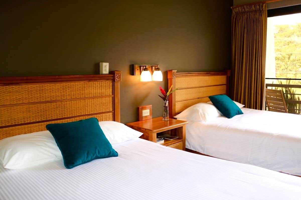 Habitación de hotel con dos camas individuales, una mesita de noche con lámpara, una pequeña mesa con un jarrón de flores y vistas al exterior a través de una ventana con cortinas.