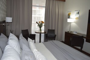 Habitación de hotel con cama, dos sillas, una mesita con un florero, un escritorio con lámpara y una obra de arte en la pared.